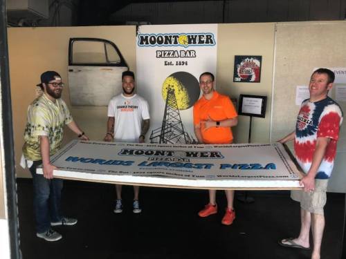 Four people hold a large banner for MoonTower Pizza Bar, promoting it as the "World's Biggest Pizza."