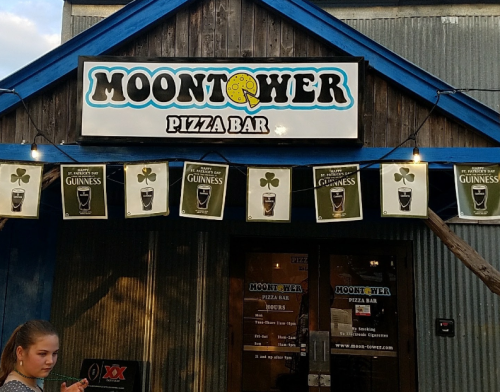 Sign for Moontower Pizza Bar with beer banners and a girl standing nearby. The building has a rustic design.