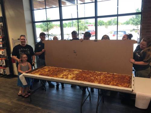 A group of people holds a giant pizza in a restaurant, with a child in front smiling at the camera.
