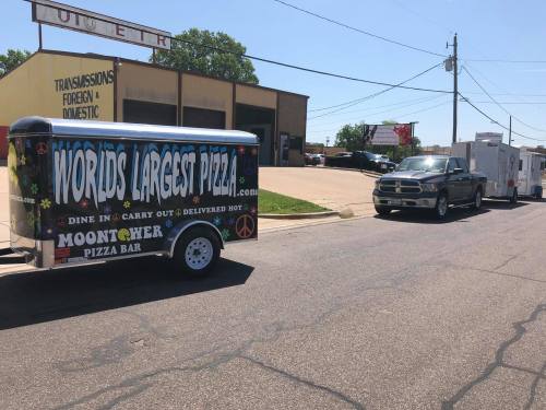 A food trailer labeled "World's Largest Pizza" parked on a sunny street, with a truck and other trailers nearby.