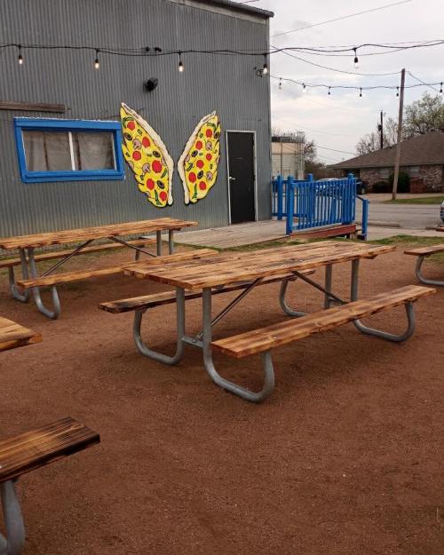 Outdoor seating area with wooden picnic tables and a colorful pizza-themed mural on a gray building.
