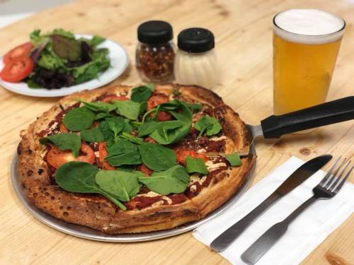 A pizza topped with spinach and tomatoes, served with a side salad and a glass of beer on a wooden table.