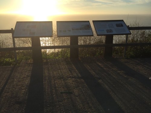 Three informational signs stand on a path overlooking the ocean at sunset, casting long shadows on the ground.