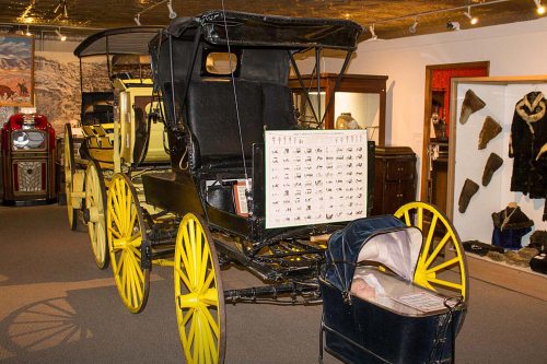 A vintage horse-drawn carriage with yellow wheels displayed in a museum, featuring a small baby carriage beside it.