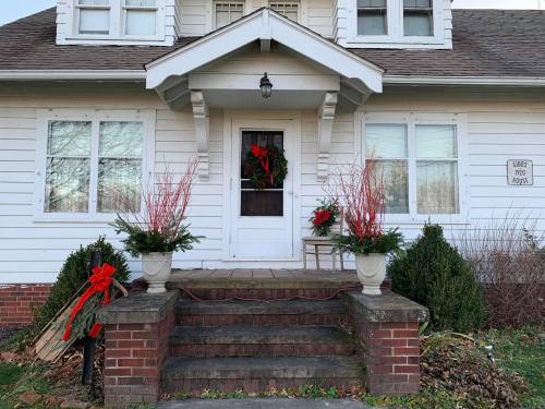 A charming white house with a decorated front porch, featuring wreaths, red bows, and potted plants.