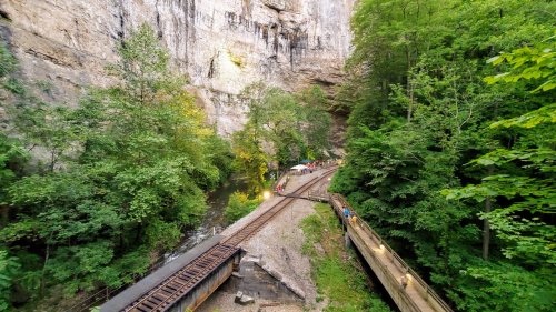 A scenic view of a rocky canyon with lush greenery, a railway track, and a wooden walkway along a stream.