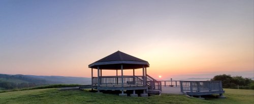 A gazebo on a grassy hill at sunrise, with soft colors in the sky and distant mountains in the background.