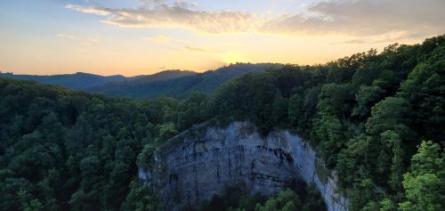 A scenic view of a cliff surrounded by lush green trees, with a sunset illuminating the sky in the background.