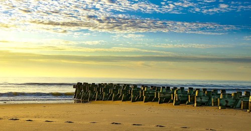 A serene beach scene at sunset, featuring wooden posts along the shore and a colorful sky with scattered clouds.