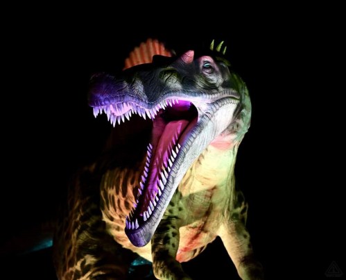 A colorful, illuminated dinosaur sculpture with an open mouth and sharp teeth against a dark background.