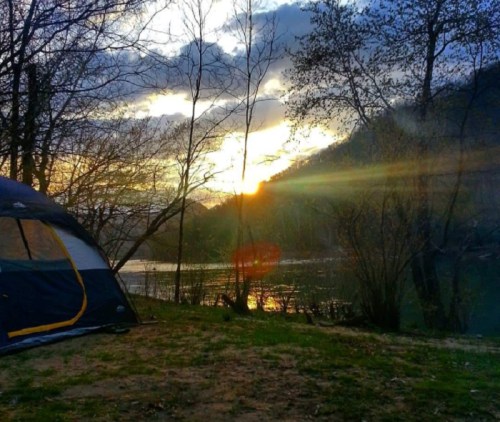 A tent by a river at sunset, with trees silhouetted against a colorful sky.