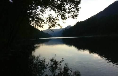 A serene river scene at dusk, with mountains in the background and trees reflecting on the water's surface.