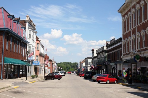 A quiet small-town street lined with shops and cars under a blue sky with fluffy clouds.