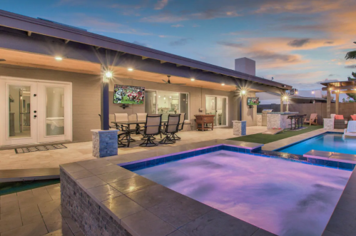 Modern backyard with a pool, hot tub, and outdoor seating, illuminated by evening lights under a colorful sky.