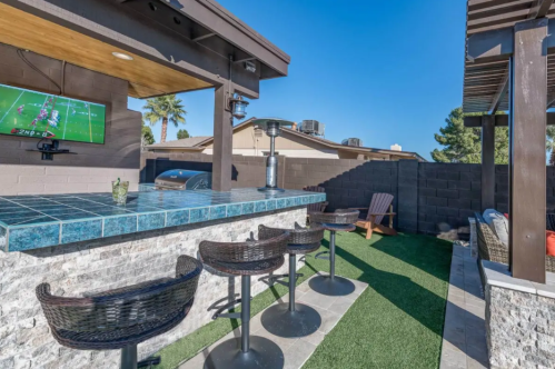 Outdoor bar area with a blue tiled counter, stools, and a TV, surrounded by green grass and a sunny sky.