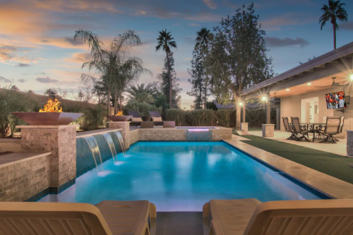 A serene backyard pool with a waterfall, surrounded by palm trees and lounge chairs, under a colorful sunset sky.