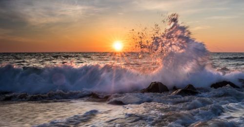 A vibrant sunrise over Ocean City Beach in MD, with waves crashing against rocks in the foreground, creating a spray of water.
