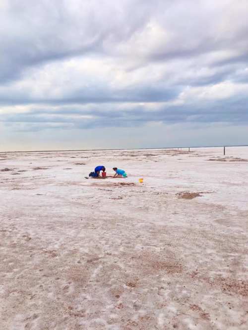 Two people crouch on a vast, dry salt flat under a cloudy sky, engaged in an activity.