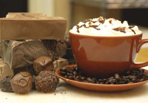 A brown cup of whipped cream topped hot chocolate, surrounded by chocolate bars, truffles, and coffee beans.
