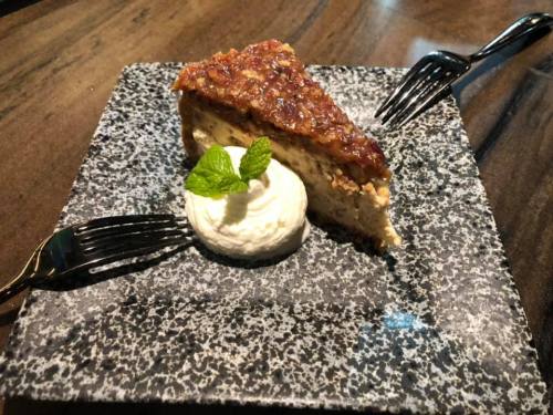 A slice of pecan pie on a textured plate, garnished with whipped cream and a mint leaf, with two forks beside it.
