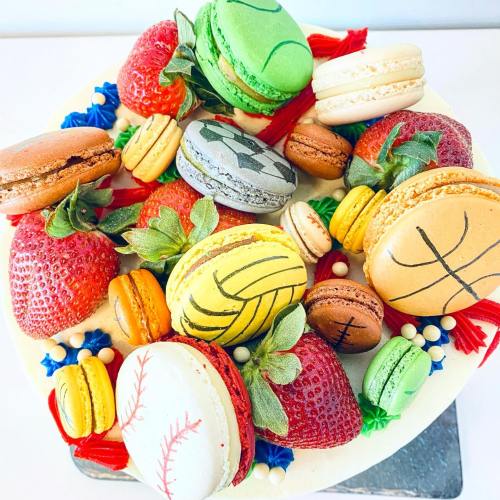 A colorful cake topped with various macarons and strawberries, featuring sports-themed designs on some macarons.