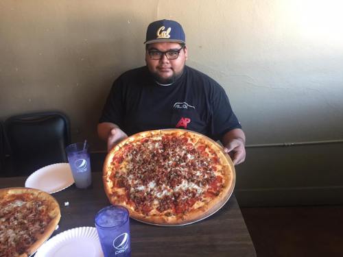 A person holding a large pizza topped with meat, sitting at a table with drinks in a casual setting.