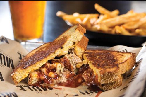 A grilled sandwich filled with pulled pork and macaroni, served with fries and a glass of beer in the background.