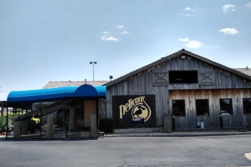 Exterior of a rustic restaurant with a blue awning and a large "Pelican" sign on a wooden building. Clear blue sky above.