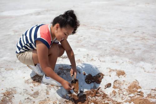 A young girl in a striped shirt digs in the ground with a small shovel on a sandy surface.