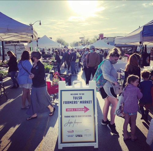 A bustling farmers' market with vendors and shoppers, tents set up, and a sign welcoming visitors to the Tulsa Farmers' Market.