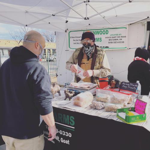 A vendor at a market selling homemade dog treats and meats, with a customer inquiring at the booth.