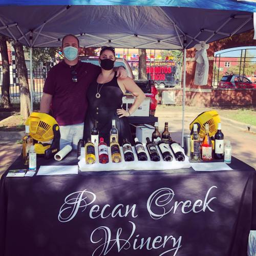 A man and woman stand behind a table with various wine bottles, under a tent labeled "Pecan Creek Winery."