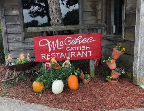 Sign for McGehee Catfish Restaurant with decorative pumpkins and flower pots in front of a rustic wooden building.