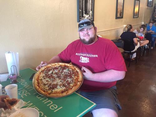A man in a red shirt holds a large pizza in a casual restaurant setting.
