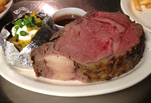 A plate featuring a large slice of prime rib, a baked potato with toppings, and a small cup of sauce.
