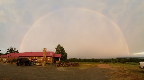 A rustic building with a red roof under a vibrant rainbow, surrounded by green fields and trees.