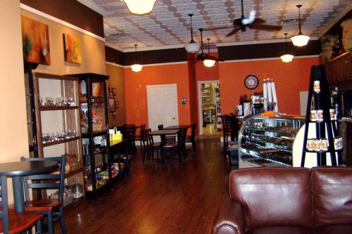 Cozy café interior with wooden floors, a display case, shelves of goods, and tables for seating. Warm, inviting atmosphere.