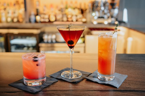 Three colorful cocktails on a bar: a pink drink, a martini with a cherry, and an orange drink, all on black napkins.