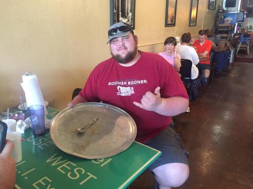 A man in a red shirt sits at a table with an empty large platter, smiling and making a hand gesture.