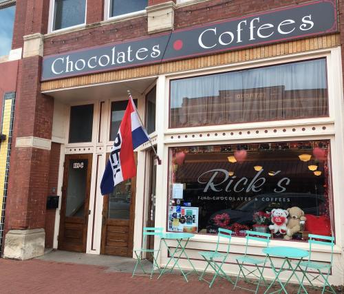 Exterior of Rick's Chocolates & Coffees, featuring a red brick facade, a flag, and green chairs outside the window.