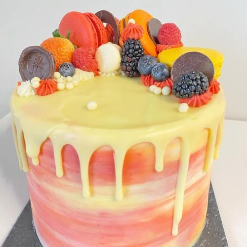 A colorful cake with a smooth drip, topped with macarons, berries, and decorative sweets.