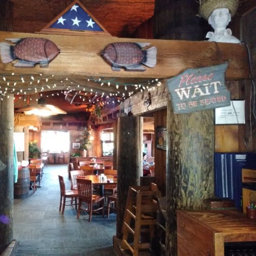 Interior of a rustic restaurant with wooden decor, fish carvings, and a sign saying "Please wait to be seated."