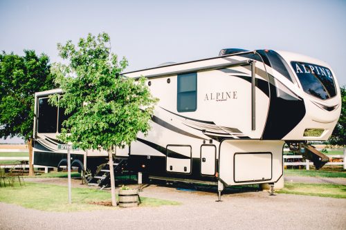 A large, modern RV parked on a grassy area, surrounded by trees and a clear blue sky.