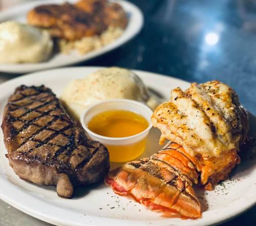 A plate featuring grilled steak, lobster tail, a small cup of butter, and mashed potatoes.