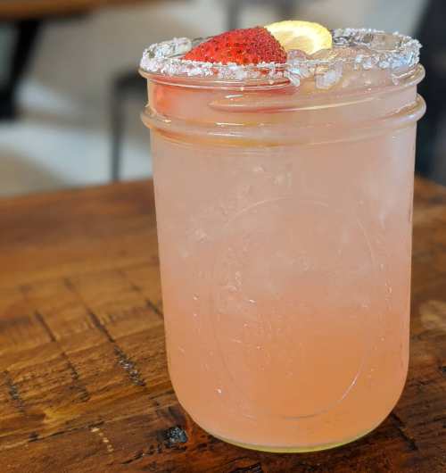 A refreshing pink drink in a mason jar, garnished with a strawberry and lemon slice, on a wooden table.