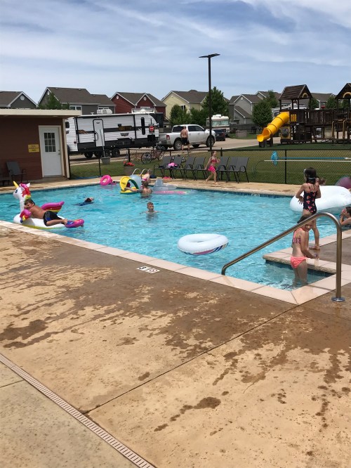 A community pool with children playing, colorful floats, and a playground in the background on a sunny day.