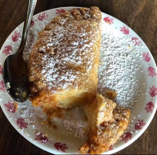 A slice of dessert dusted with powdered sugar on a decorative plate, with a spoon resting beside it.