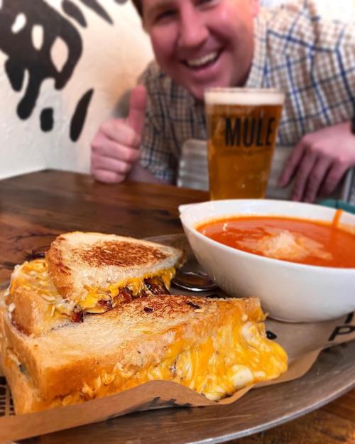 A grilled cheese sandwich with melted cheese, a bowl of tomato soup, and a beer, with a smiling person in the background.
