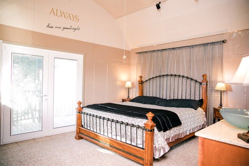 Cozy bedroom featuring a wooden bed, soft bedding, and large windows with sheer curtains. Wall decor includes a quote.