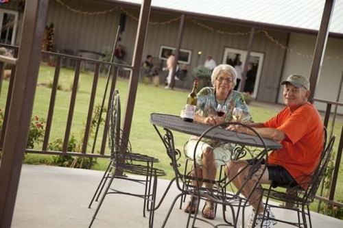 An elderly couple sits at a table outdoors, enjoying drinks and smiling, with a grassy area and building in the background.